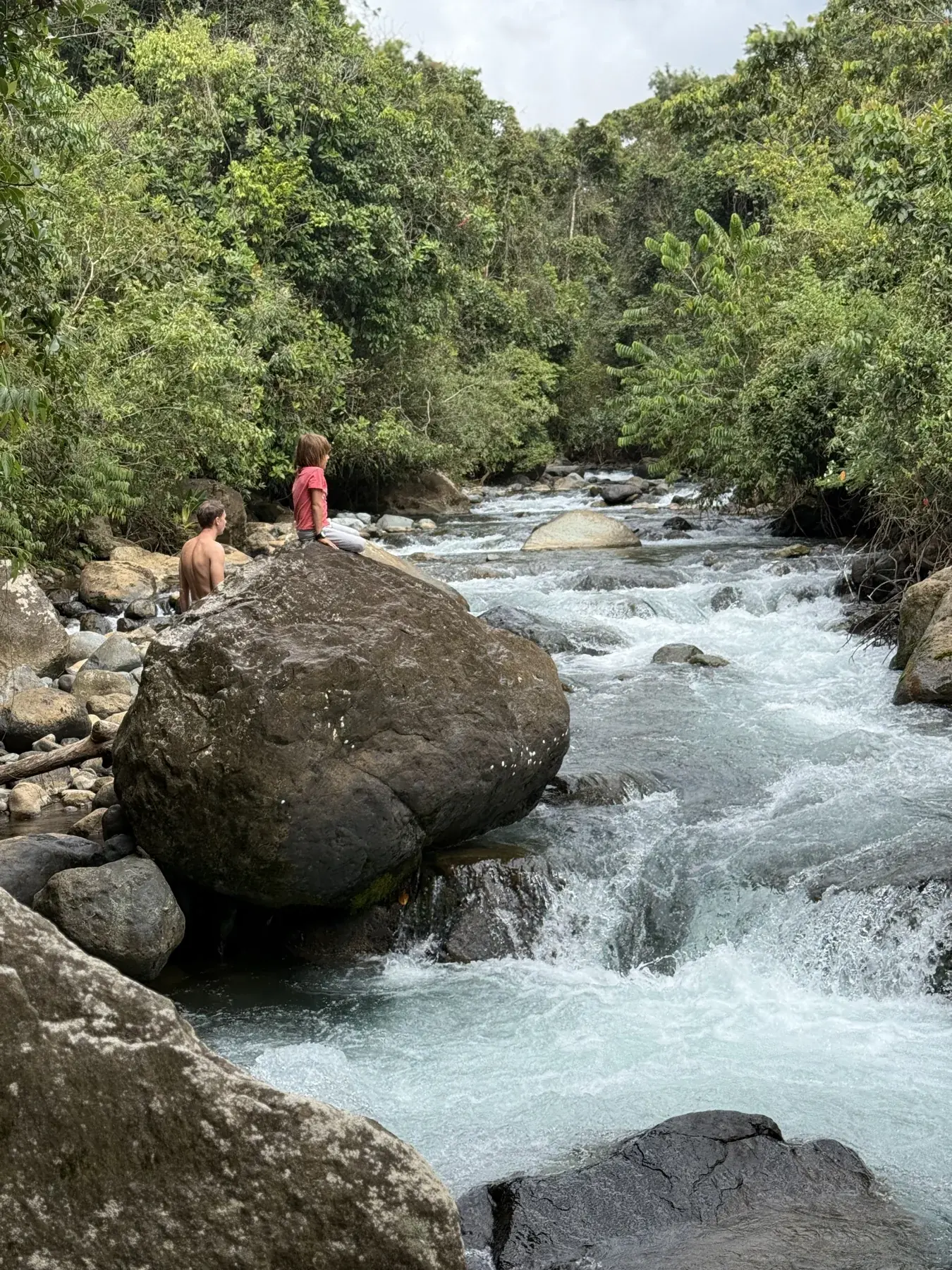 People resting on a large river boulder beside flowing water and forest
